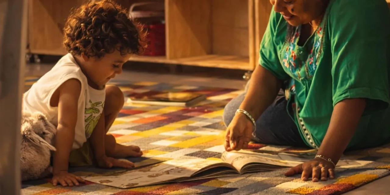 Criança folheando um livro no tapete colorido, ao lado de uma educadora
